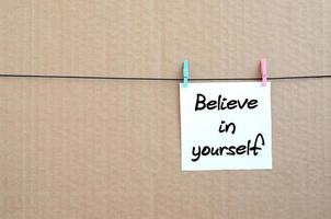 Believe in yourself. Note is written on a white sticker that hangs with a clothespin on a rope on a background of brown cardboard photo