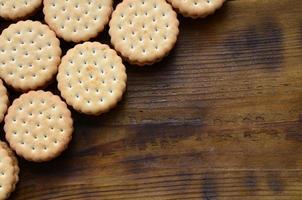 A round sandwich cookie with coconut filling lies in large quantities on a brown wooden surface. Photo of edible treats on a wooden background with copy space