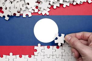 Laos flag  is depicted on a table on which the human hand folds a puzzle of white color photo