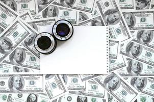 dos lentes fotográficos y un cuaderno blanco se encuentran en el fondo de muchos billetes de dólar. espacio para texto foto