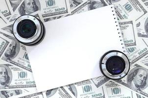 Two photographic lenses and white notebook lie on the background of a lot of dollar bills. Space for text photo