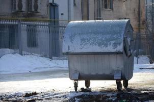 A silver garbage container stands near residential buildings in winter photo