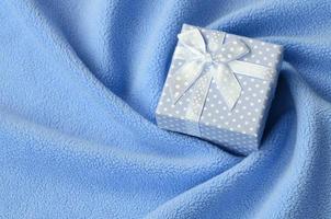A small gift box in blue with a small bow lies on a blanket of soft and furry light blue fleece fabric with a lot of relief folds. Packing for a gift to your lovely girlfriend photo