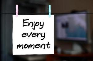 Enjoy every moment. Note is written on a white sticker that hangs with a clothespin on a rope on a background of office interior photo