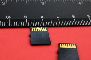 Two small micro SD memory cards lie on a red background next to a black ruler. A small and compact data and information store photo