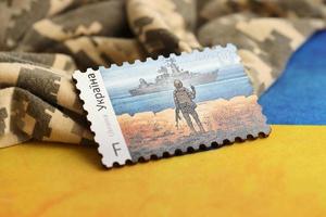 TERNOPIL, UKRAINE - SEPTEMBER 2, 2022 Famous Ukrainian postmark with russian warship and ukrainian soldier as wooden souvenir on army camouflage uniform and national flag photo