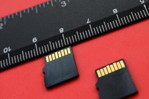 Two small micro SD memory cards lie on a red background next to a black ruler. A small and compact data and information store photo