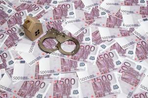 Scam with real estate. Cheating when buying or renting a house. Punishment for deception. A toy house with handcuffs on a many 500 purple euro banknotes photo