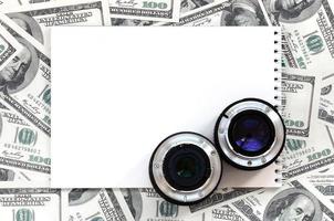 Two photographic lenses and white notebook lie on the background of a lot of dollar bills. Space for text photo