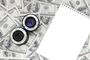 Two photographic lenses and white notebook lie on the background of a lot of dollar bills. Space for text photo