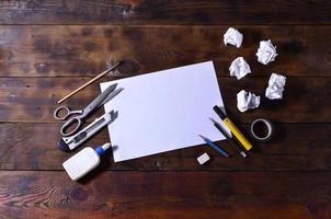 A school or office still life with a white blank sheet of paper and many office supplies. The school supplies lie on a brown wooden background. Place for text photo