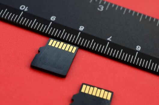 Two small micro SD memory cards lie on a red background next to a black ruler. A small and compact data and information store photo
