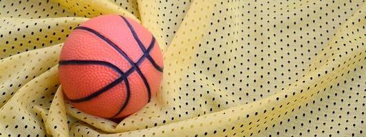 Small orange rubber basketball lies on a yellow sport jersey clothing fabric texture and background with many folds photo