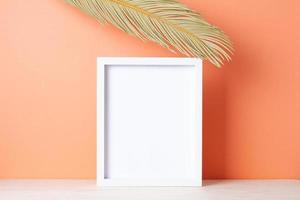 Home decoration with white mock up frame and palm leaf on table. Artwork showcase. Close up, copy space photo
