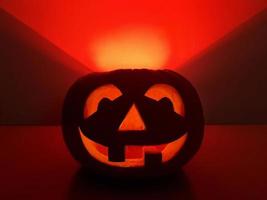 Halloween holiday background with orange smiling jack-o-lantern. Big carved glowing pumpkin stands centered on the surface in dark night. The back wall highlighted in red triangle over pumpkin. photo