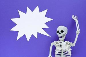 Skeleton on violet background with white blank paper thorny sound bubble. Anatomical plastic model human skeleton with one raised hand screams loudly. Empty yell cloud, copy space. Purple Halloween. photo