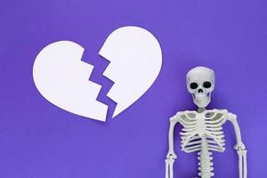 Skeleton on violet background with white blank paper cut big broken heart. Anatomical plastic model human skeleton on purple color background. Valentine's day concept. photo