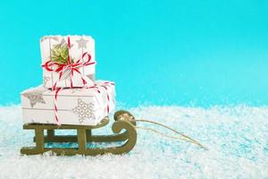 Two gift boxes wrapped of white and gray paper with red-white ribbon, decorated of fir branch on a green wooden sleigh on a blue background with artificial snow. Christmas and New Year concept. photo