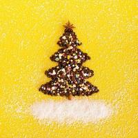 árbol de navidad hecho de granos de café y estrella de anís decorada, aspersión culinaria multicolor y chips de coco sobre un fondo amarillo, vista superior. foto