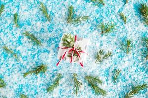 Small gift box wrapped of white and gray paper with red-white ribbon on the blue background with artificial snow decorated fir branches, top view. Christmas and New Year concept with copy space. photo