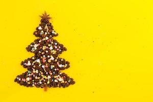 Winter composition with Christmas tree made by coffee beans and decorated anise star, cinnamon stick and multicolored culinary sprinkling on a yellow background, flat lay. Greeting card for New Year. photo