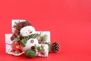 Christmas still life with snowman and gift boxes wrapped of white-gray paper with red-white ribbons on a red background, decorated of fir branches, pine cone and red wooden decorations. photo