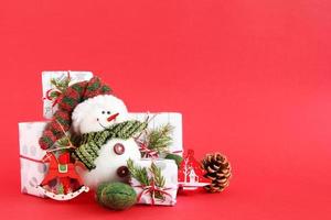 Christmas still life with snowman and gift boxes wrapped of white-gray paper with red-white ribbons on a red background, decorated of fir branches, pine cone and red wooden decorations. photo