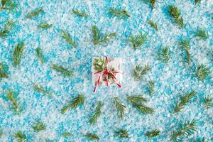 Small gift box wrapped of white and gray paper with red-white ribbon on the blue background with artificial snow decorated fir branches, top view. Christmas and New Year concept with copy space. photo