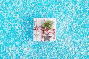 Gift box wrapped of white and gray paper, decorated red-white ribbon and fir branch on the blue background with artificial snow, top view. Christmas and New Year concept with copy space. photo