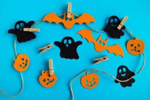 Preparation for Halloween. Black and orange paper decorations - ghosts, pumpkins and bats on a rope with pins on a blue background, top view. photo