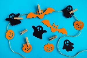 Preparation for Halloween. Black and orange paper decorations - ghosts, pumpkins and bats on a rope with pins on a blue background, top view. photo
