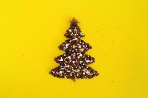 Winter composition with Christmas tree made by coffee beans and decorated anise star, cinnamon stick and multicolored culinary sprinkling on a yellow background, flat lay. Greeting card for New Year. photo