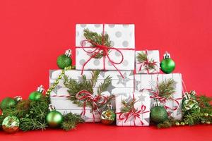 cajas de regalo de navidad envueltas en papel blanco-gris con cintas rojo-blanco sobre un fondo rojo, decoradas con ramas de abeto y adornos navideños de color verde brillante. concepto de navidad y año nuevo. foto