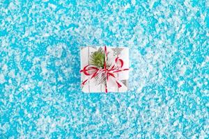 Gift box wrapped of white and gray paper, decorated red-white ribbon and fir branch on the blue background with artificial snow, top view. Christmas and New Year concept with copy space. photo