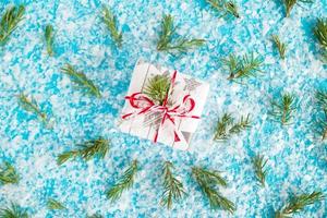 Small gift box wrapped of white and gray paper with red-white ribbon on the blue background with artificial snow decorated fir branches, top view. Christmas and New Year concept with copy space. photo