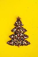 Winter composition with Christmas tree made by coffee beans and decorated anise star, cinnamon stick and multicolored culinary sprinkling on a yellow background, flat lay. Greeting card for New Year. photo