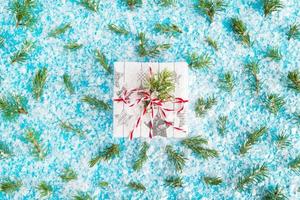 Gift box wrapped of white and gray paper with red-white ribbon on the blue background with artificial snow decorated fir branches, top view. Christmas and New Year concept with copy space. photo