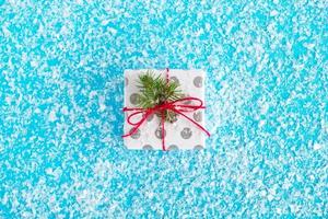 Gift box wrapped of white and gray paper, decorated red-white ribbon and fir branch on the blue background with artificial snow, top view. Christmas and New Year concept with copy space. photo