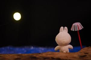 A rabbit doll standing besides an umbrella looking out to the blue sea on the night with the moon light. photo
