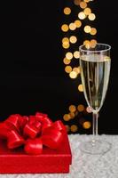 Red christmas gift box with blurred focus of a glass of sparkling wine and golden bokeh lights on dark background. photo