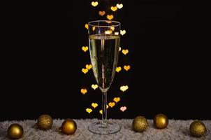 una copa de vino espumoso con adornos navideños dorados con luces bokeh en forma de amor sobre fondo oscuro. foto