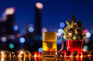 un vaso de cerveza se pone sobre una mesa de madera que tiene un árbol de navidad y adornos de adorno con un colorido fondo de luz de la ciudad. foto