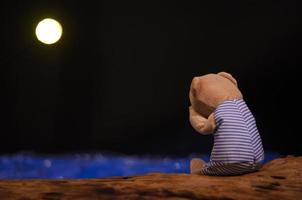 oso de peluche llorando y sentado solo en una madera frente al mar azul y la luna en la noche de tristeza. foto