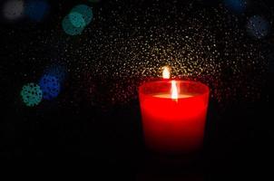 Burning candle in red glass puts beside window with rain drop on bokeh light and dark background. photo