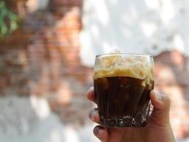 la mano sosteniendo un vaso de espresso agitado mezclando cubitos de hielo con café para una bebida fría para disfrutar del clima cálido del verano con el fondo de la vieja pared de ladrillo foto