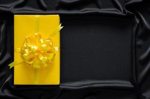 A yellow gift box with ribbon puts on black smooth and wavy cloth with space for text. Black friday and Boxing day concept. photo