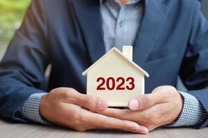 Businessman hands holding wooden House model with 2023 New Year text. Property insurance and real estate concepts photo