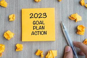 2023 Goal, Plan, Action word on yellow note with Businessman holding pen and crumbled paper on wooden table background. New Year New Start, Resolutions, Strategy  concept photo