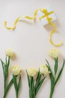 Gift box wrapped in white paper with a yellow ribbon and flowers of yellow tulips on a light table. Flat lay. Copy space for text. photo