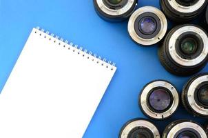 varias lentes fotográficas y un cuaderno blanco yacen sobre un fondo azul brillante. espacio para texto foto